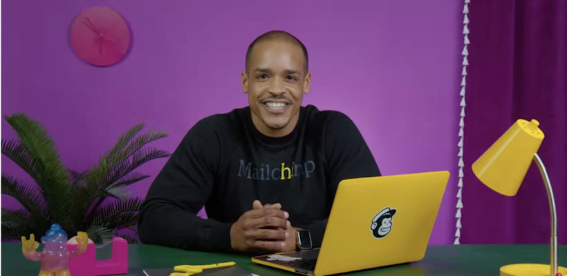 Man with shave head and black Mailchimp shirt smiling at a desk facing an open laptop