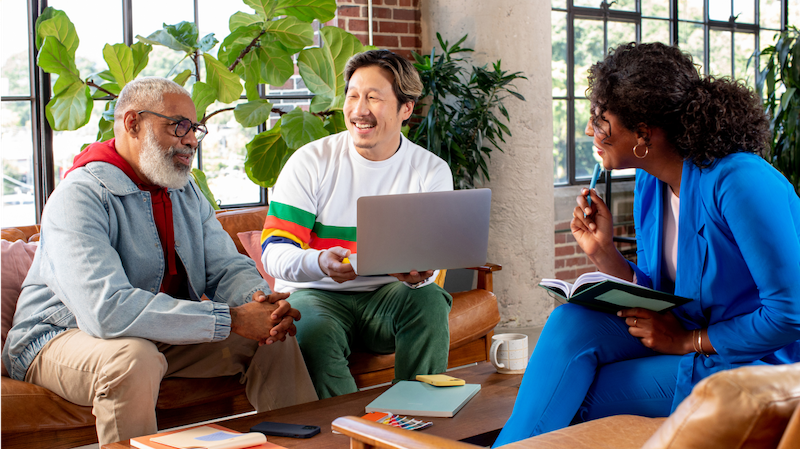 Three coworkers sit across from each other. The one in the middle holds a laptop, and the one on the right holds a book and a pen.